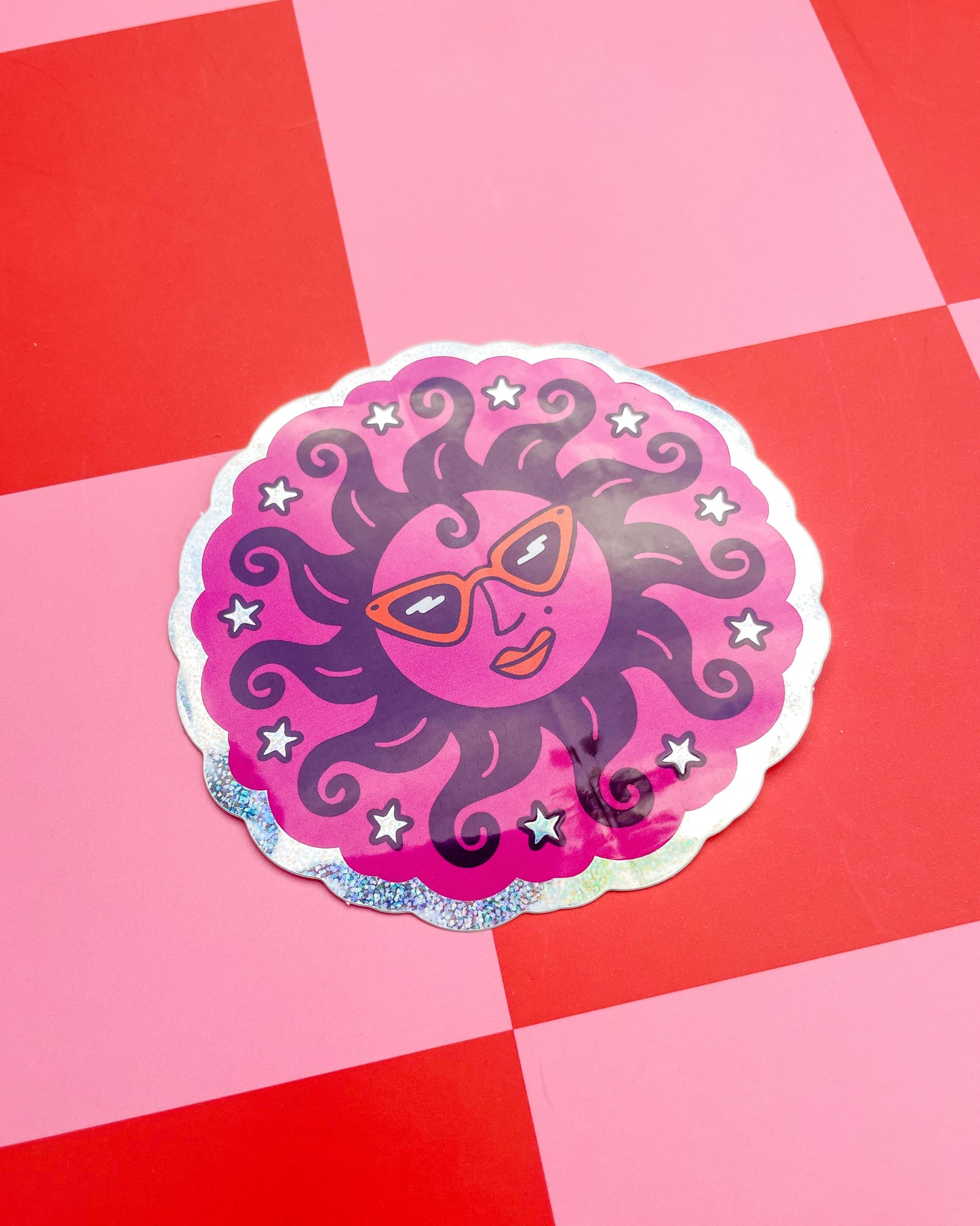 A glitter holographic scalloped circular sticker featuring a stylized sun illustration wearing orange retro sunglasses, orange lipstick, curly like hair, surrounded by stars, set against a pink and red checkered background.