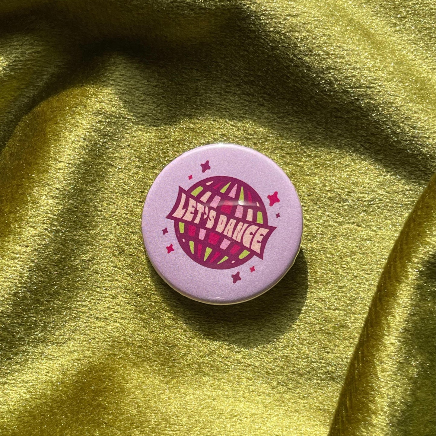 Purple pinback button with a disco ball and 'Let's Dance' text on a green fabric background