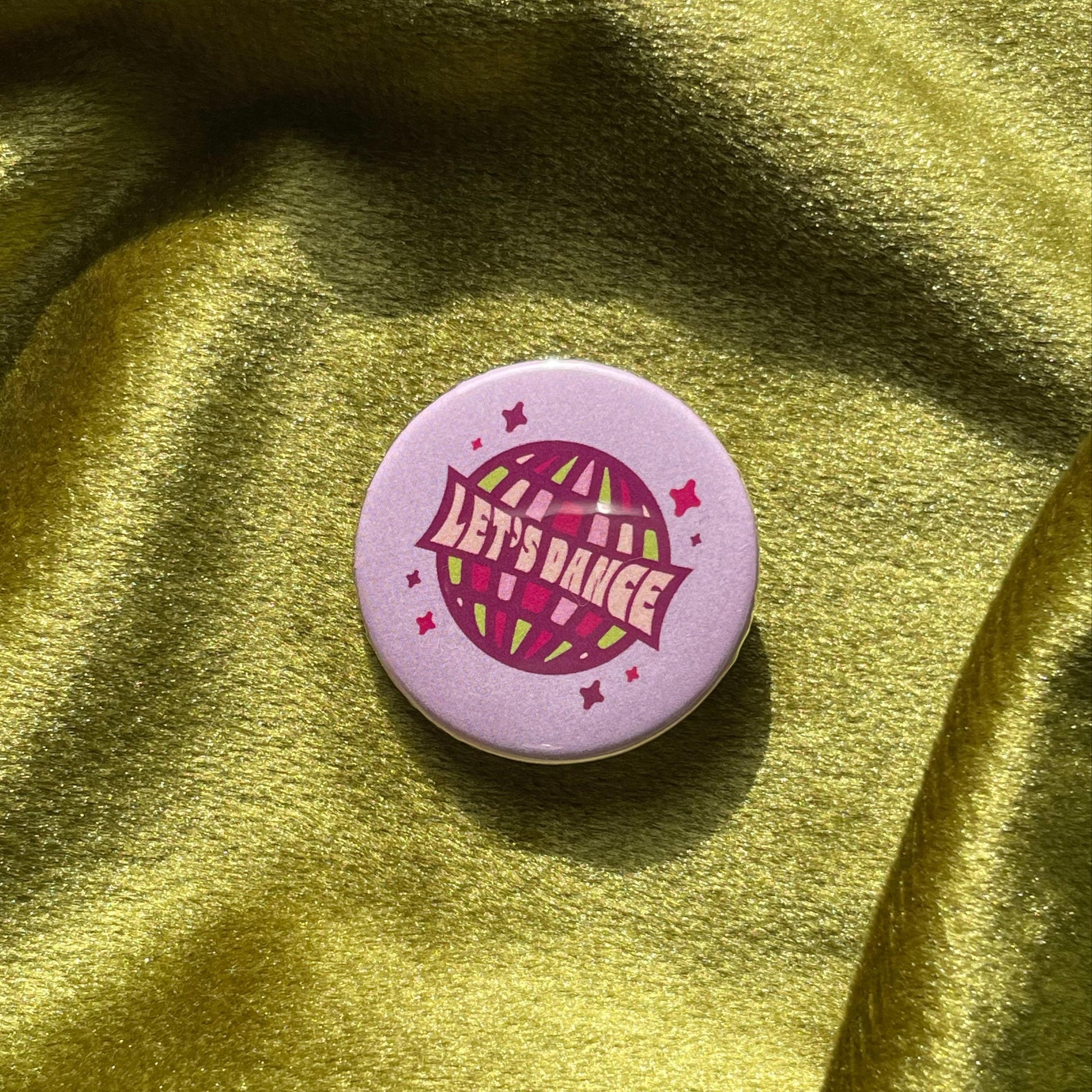 Purple pinback button with a disco ball and 'Let's Dance' text on a green fabric background