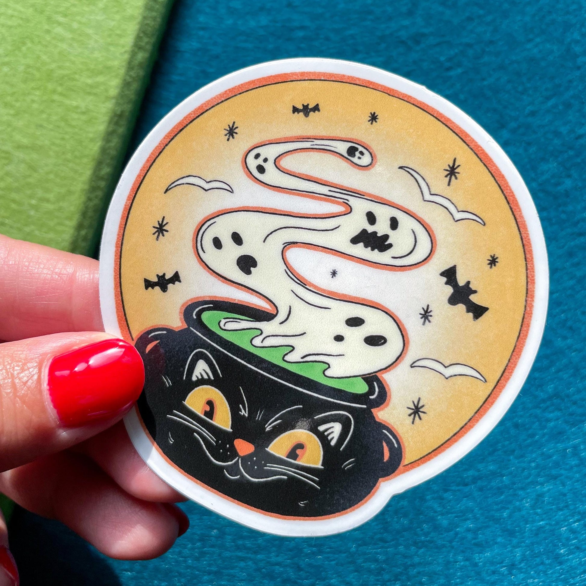 Halloween-themed sticker held by a hand with red nail polish on a blue and green background