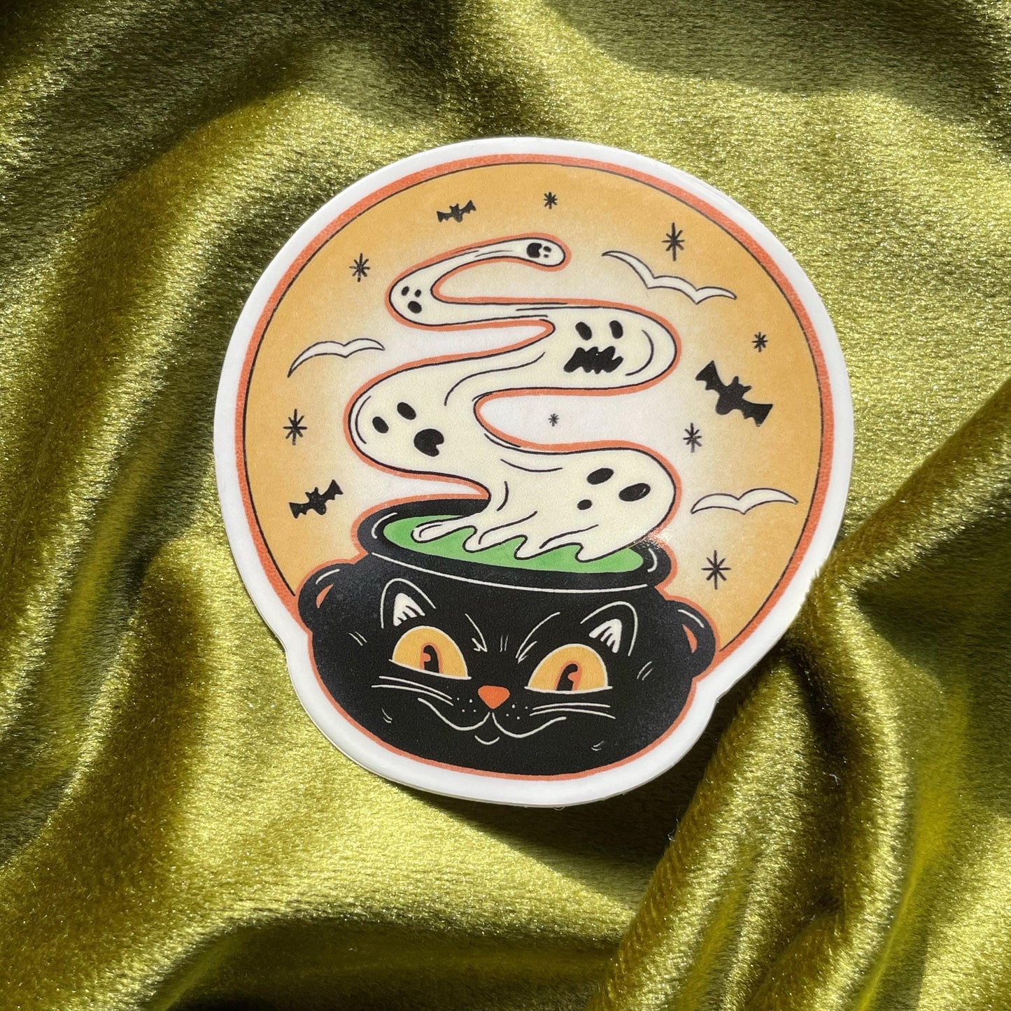 A black cat with yellow eyes peeking out of a cauldron with a white ghost rising from the green liquid inside. The cauldron is on a yellow background with bats and stars.