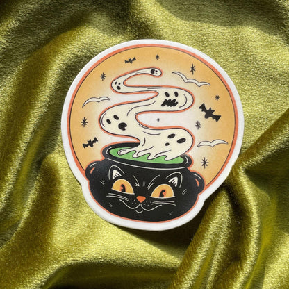 A black cat with yellow eyes peeking out of a cauldron with a white ghost rising from the green liquid inside. The cauldron is on a yellow background with bats and stars.