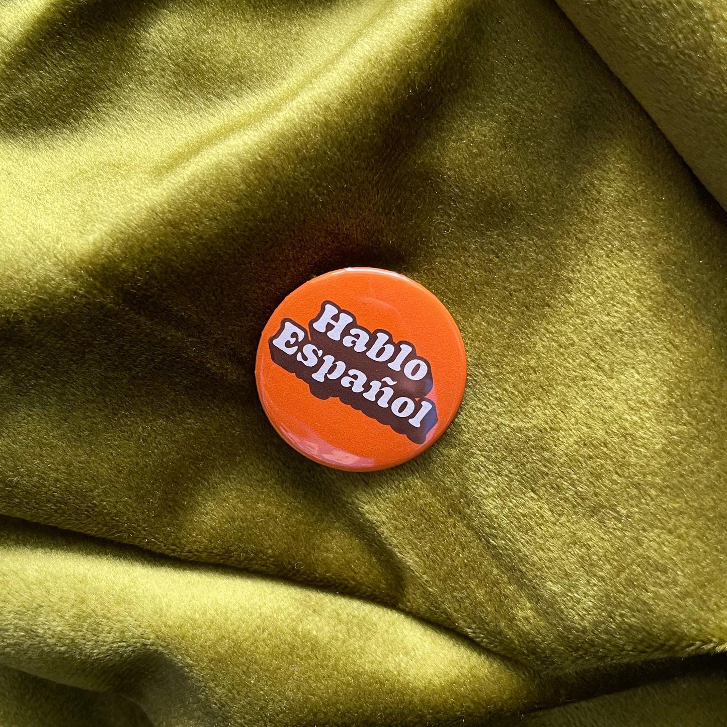 An orange button with the words 'Hablo Español' in white text. The button is round and has a glossy finish. The background is a textured, olive-green fabric.