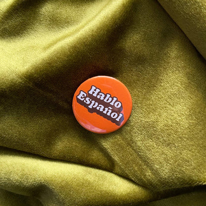 An orange button with the words 'Hablo Español' in white text. The button is round and has a glossy finish. The background is a textured, olive-green fabric.