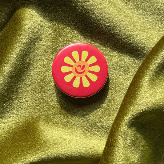 Red pinback button with yellow sunflower winking design on a green fabric background