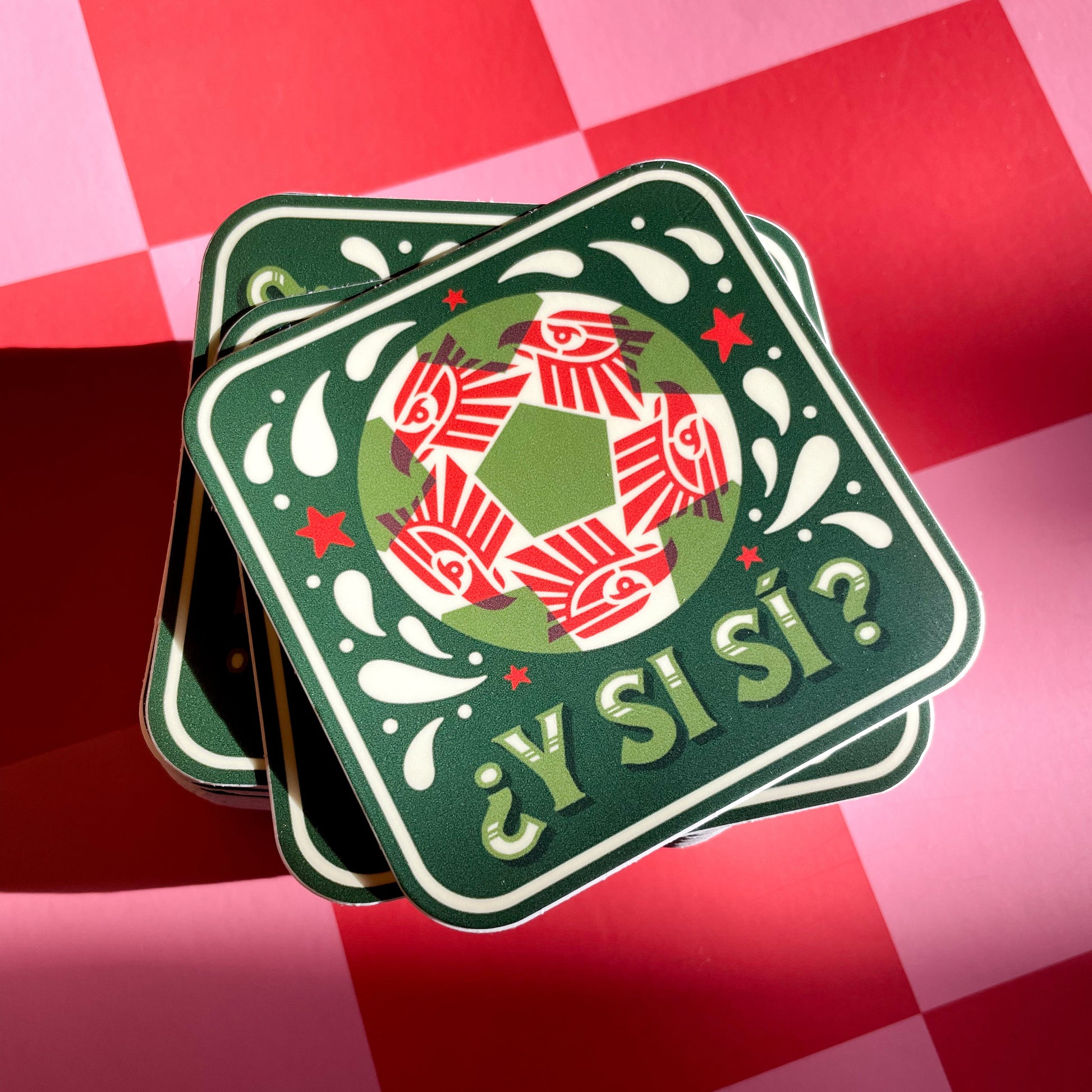 A square sticker with a red, white, and green soccer ball. A red geometric eagle pattern inside the ball, and the Spanish text '¿Y Si Sí?' in green, displayed next to a amber clover-shaped glass tray on a pink and red checkered background.