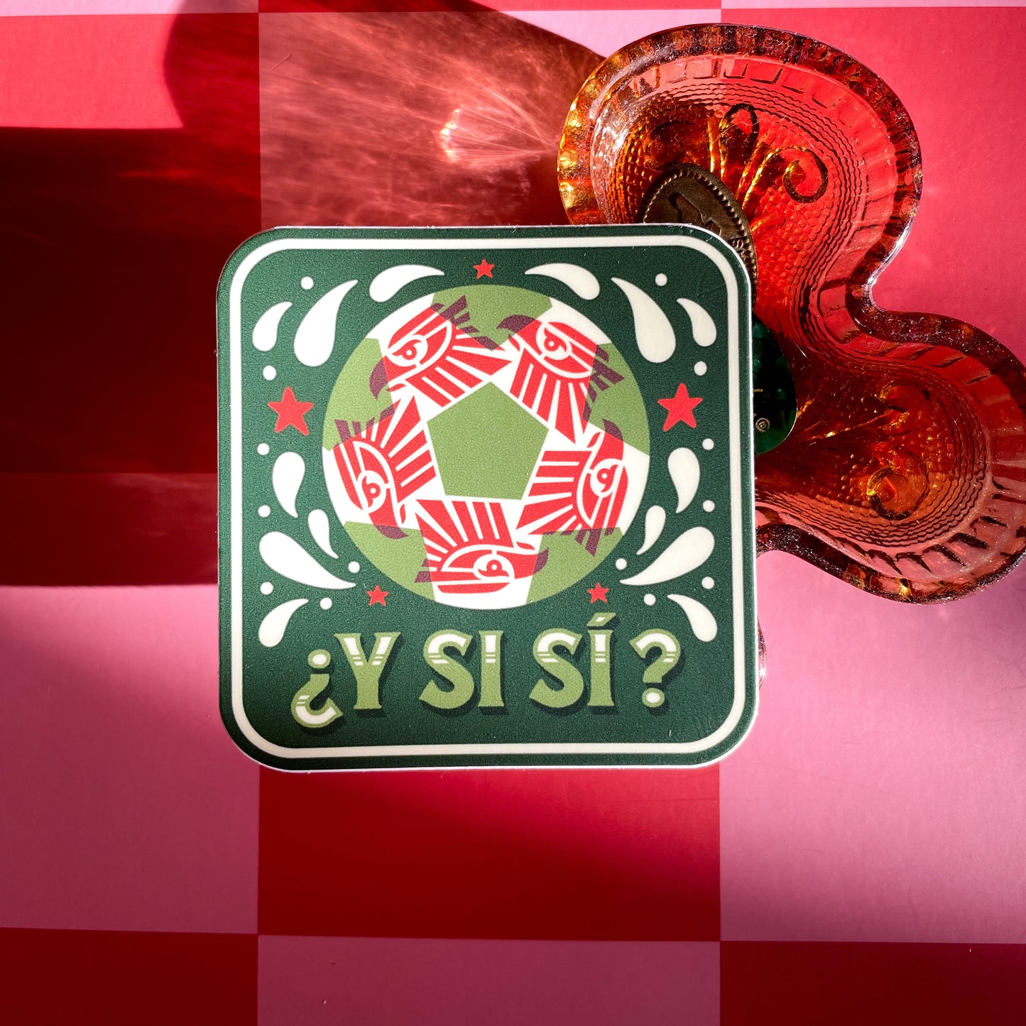A square sticker with a red, white, and green soccer ball. A red geometric eagle pattern inside the ball, and the Spanish text '¿Y Si Sí?' in green, displayed next to a amber clover-shaped glass tray on a pink and red checkered background.
