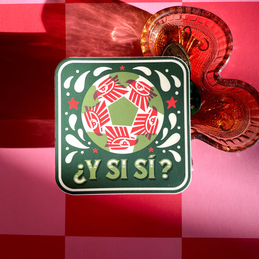 A square sticker with a red, white, and green soccer ball. A red geometric eagle pattern inside the ball, and the Spanish text '¿Y Si Sí?' in green, displayed next to a amber clover-shaped glass tray on a pink and red checkered background.