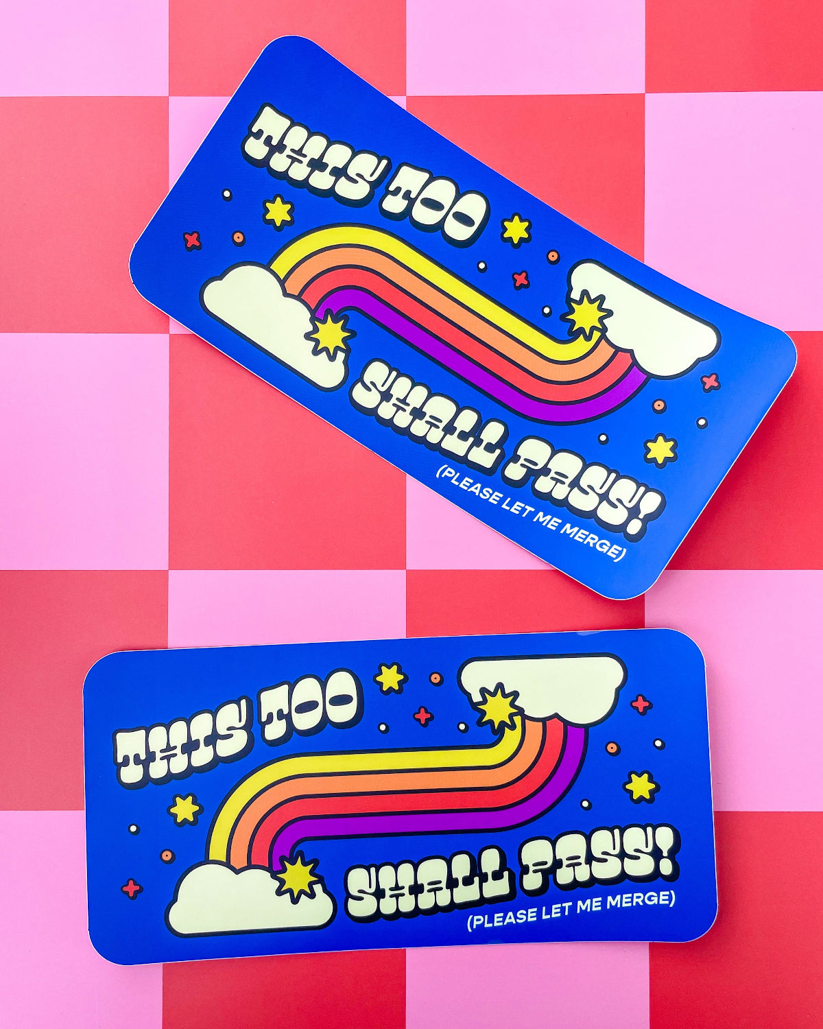Two matching blue bumper stickers with a retro rainbow design and the text 'THIS TOO SHALL PASS! (PLEASE LET ME MERGE)' are arranged on a pink and red checkered background.