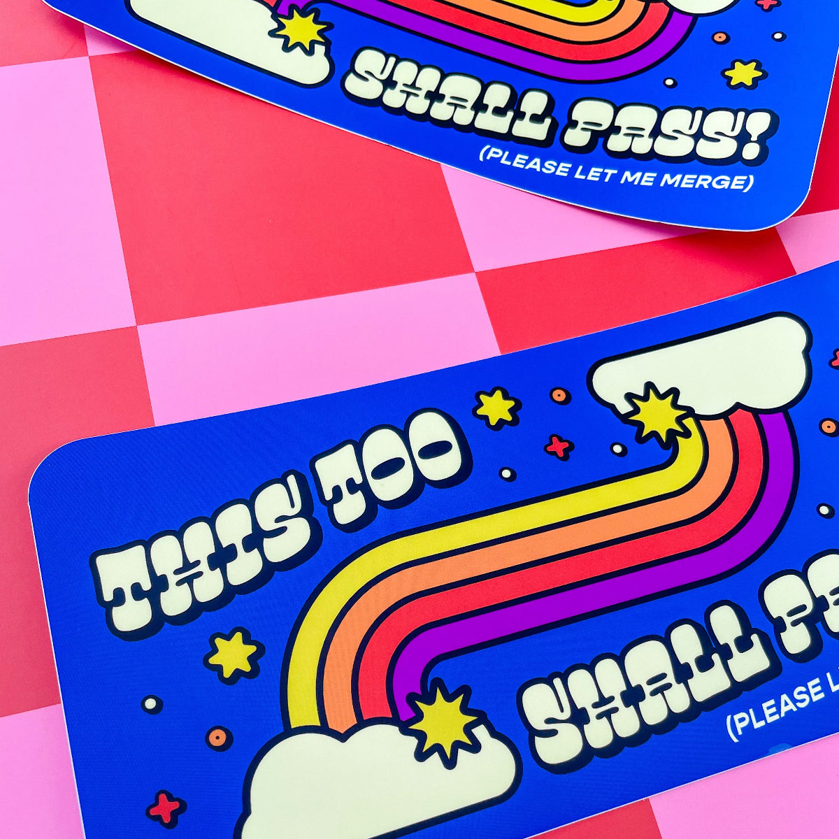 Two rectangular blue bumper stickers with a colorful rainbow, clouds, stars, and the text 'THIS TOO SHALL PASS! (PLEASE LET ME MERGE)' arranged on a pink and red checkered background.