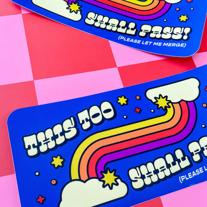Two rectangular blue bumper stickers with a colorful rainbow, clouds, stars, and the text 'THIS TOO SHALL PASS! (PLEASE LET ME MERGE)' arranged on a pink and red checkered background.