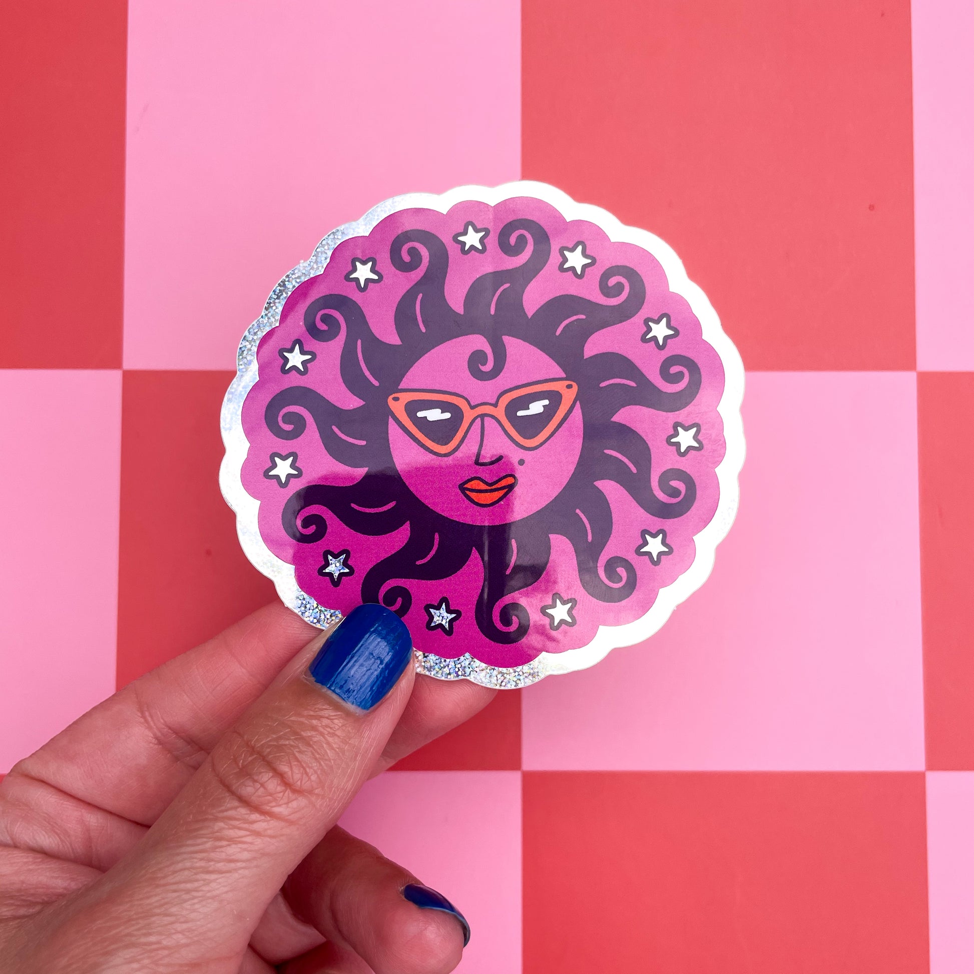A hand with blue nail polish holds a glitter holographic scalloped circular sticker featuring a stylized sun illustration wearing orange retro sunglasses, orange lipstick, curly like hair, surrounded by stars, set against a pink and red checkered background.