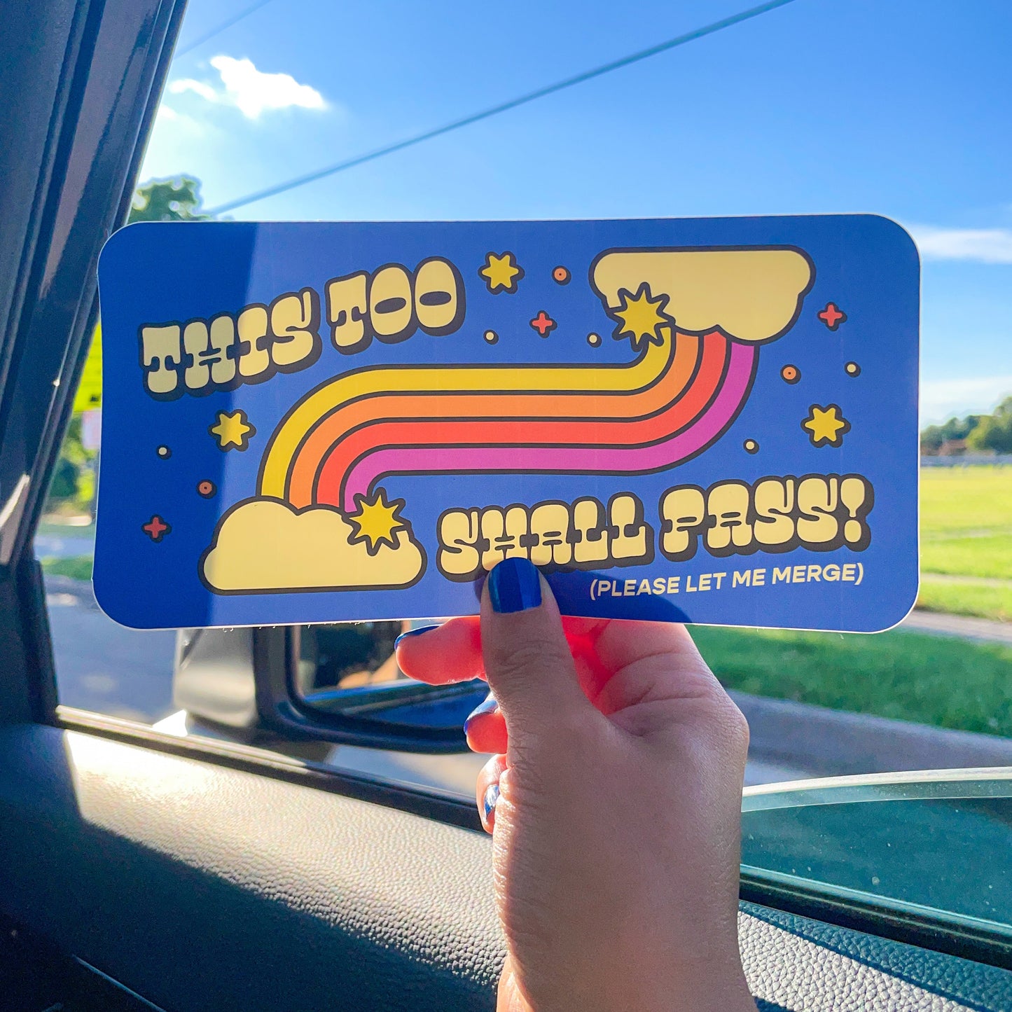 A hand holds a blue rectangular sticker with a retro rainbow design and the text 'THIS TOO SHALL PASS! (PLEASE LET ME MERGE)' inside a car window.