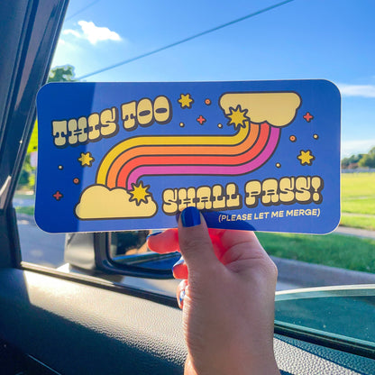 A hand holds a blue rectangular sticker with a retro rainbow design and the text 'THIS TOO SHALL PASS! (PLEASE LET ME MERGE)' inside a car window.