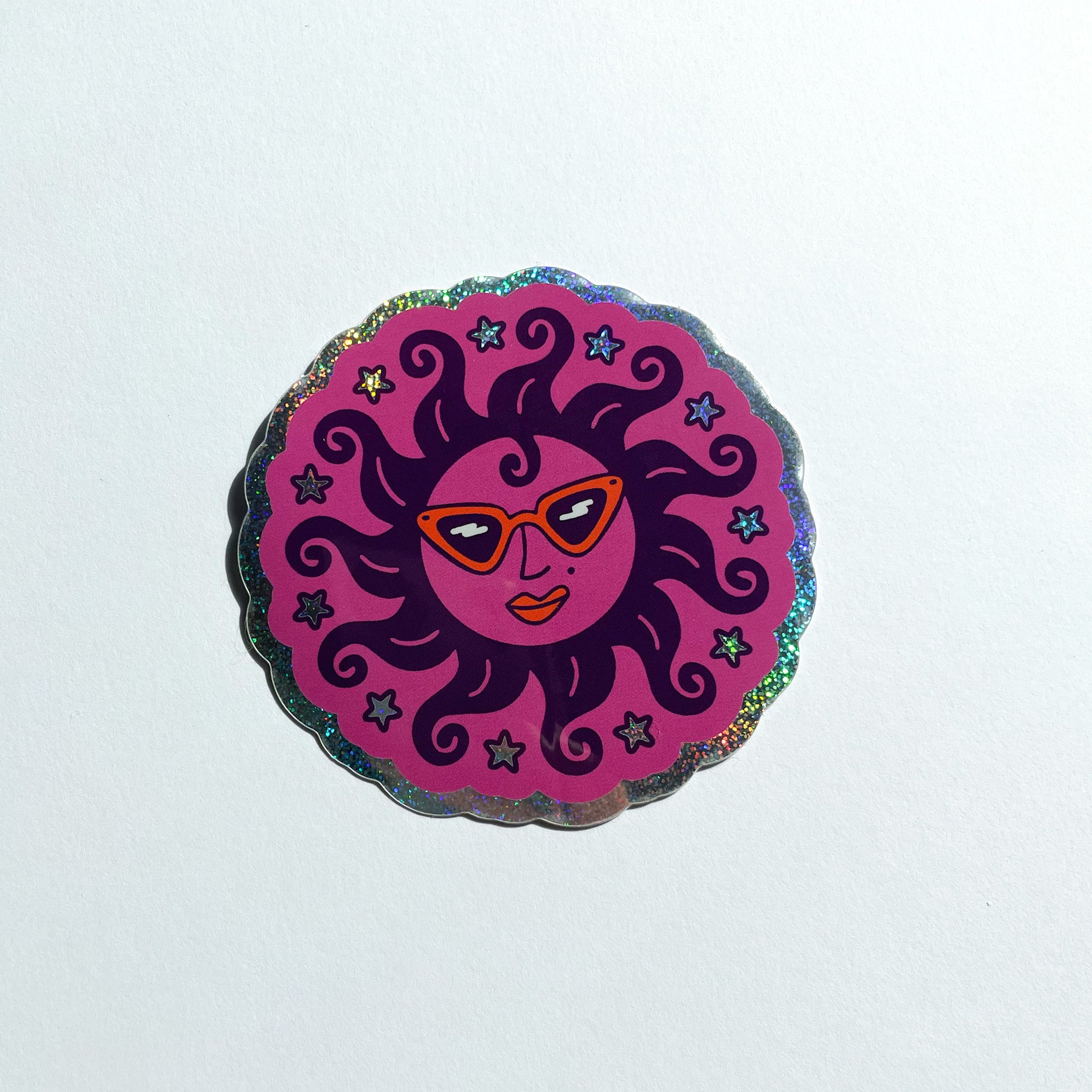 A glitter holographic scalloped circular sticker featuring a stylized sun illustration wearing orange retro sunglasses, orange lipstick, curly like hair, surrounded by stars, set against a white background.