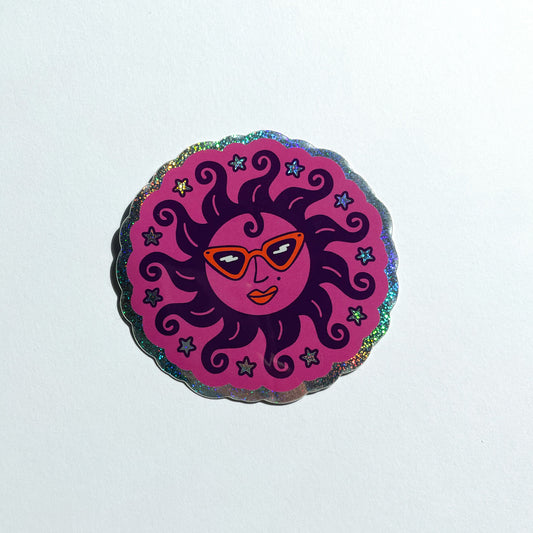 A glitter holographic scalloped circular sticker featuring a stylized sun illustration wearing orange retro sunglasses, orange lipstick, curly like hair, surrounded by stars, set against a white background.