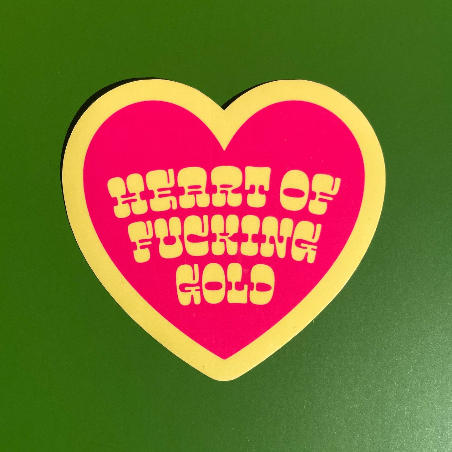 Heart-shaped sticker with yellow text on a pink background against a green background