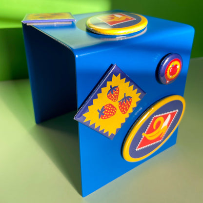 Set of three colorful magnets featuring a trio of strawberries, a peeling banana, and an apple design hanging on a blue metal stand on a green background