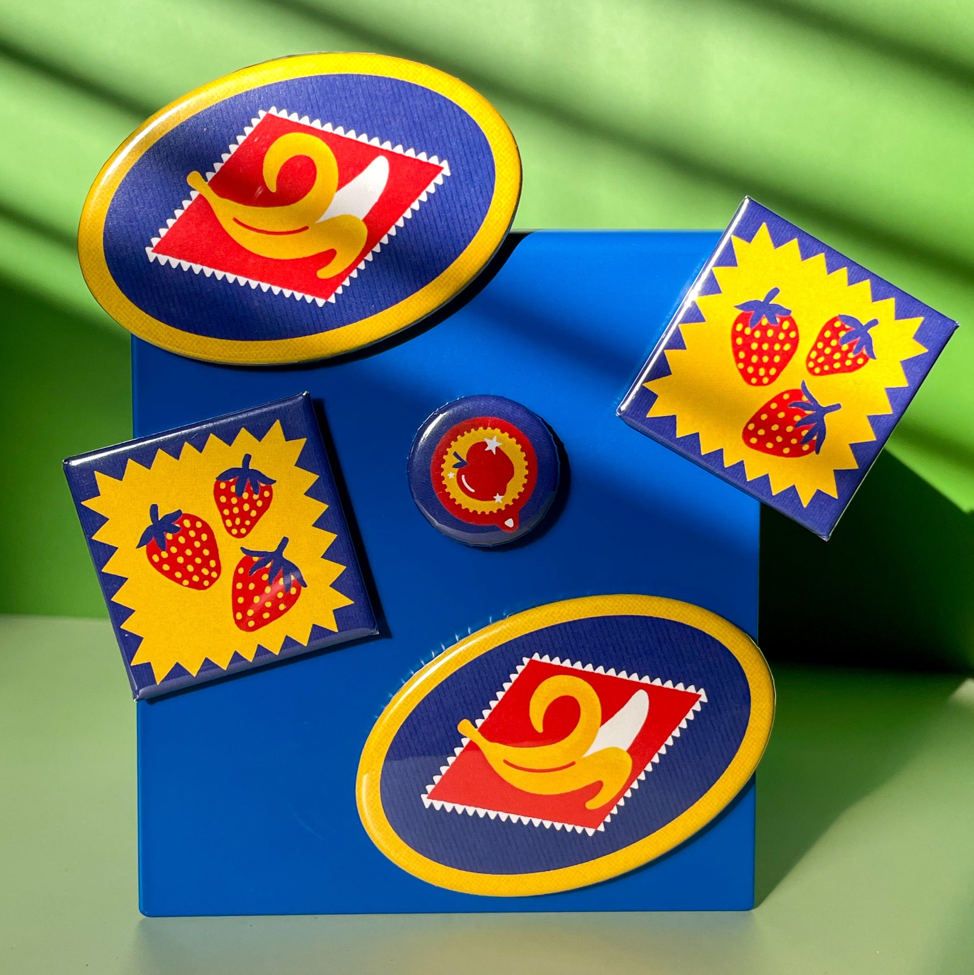 Set of three colorful magnets featuring a trio of strawberries, a peeling banana, and an apple design hanging on a blue metal stand on a green background