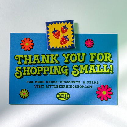 Blue postcard with colorful graphics and text on a blue background being held up by a square magnet featuring three strawberries on a yellow zig zag background with a blue border