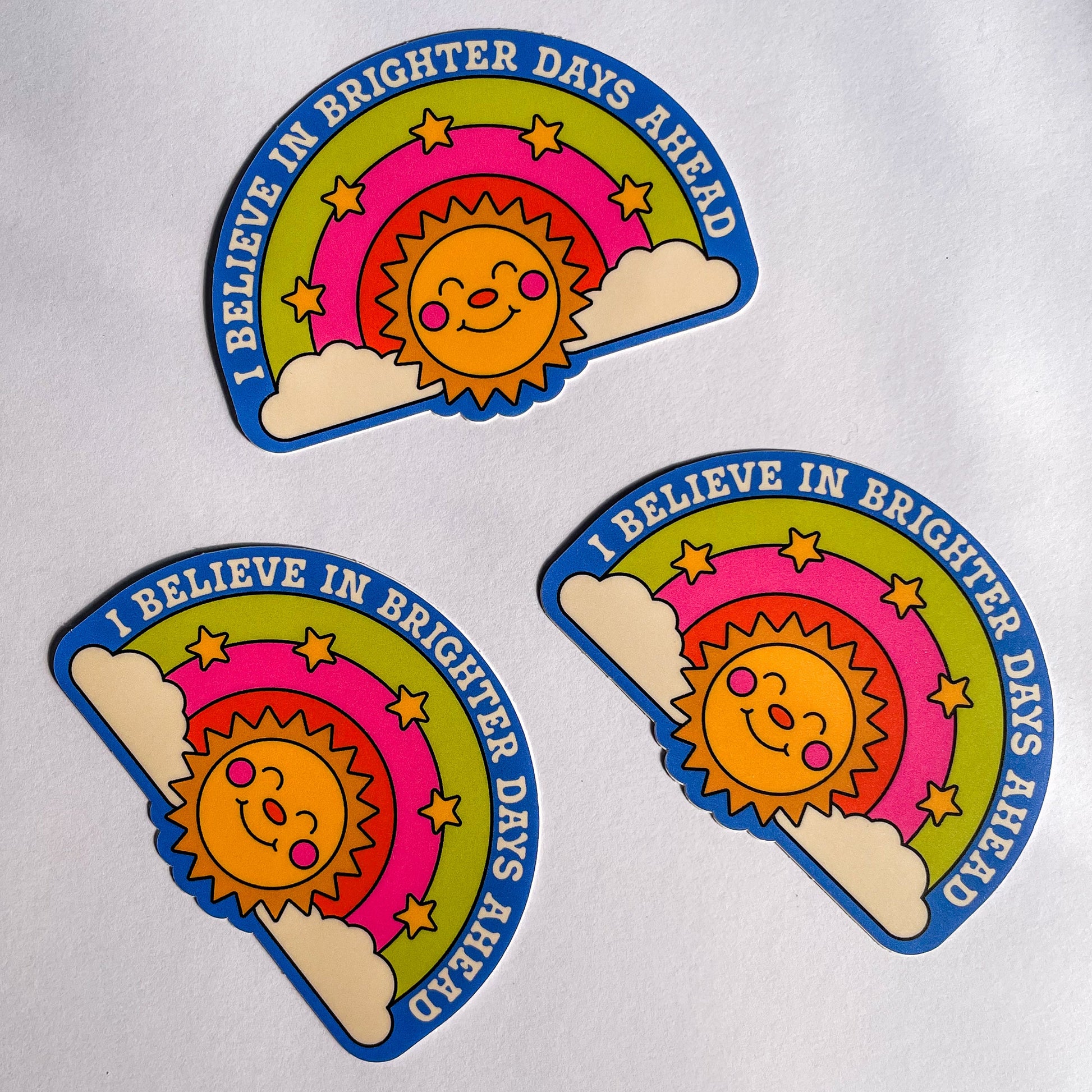 Three stickers on top of a white background featuring a smiling sun below a three color rainbow with stars and the text ' i believe in brighter days ahead'