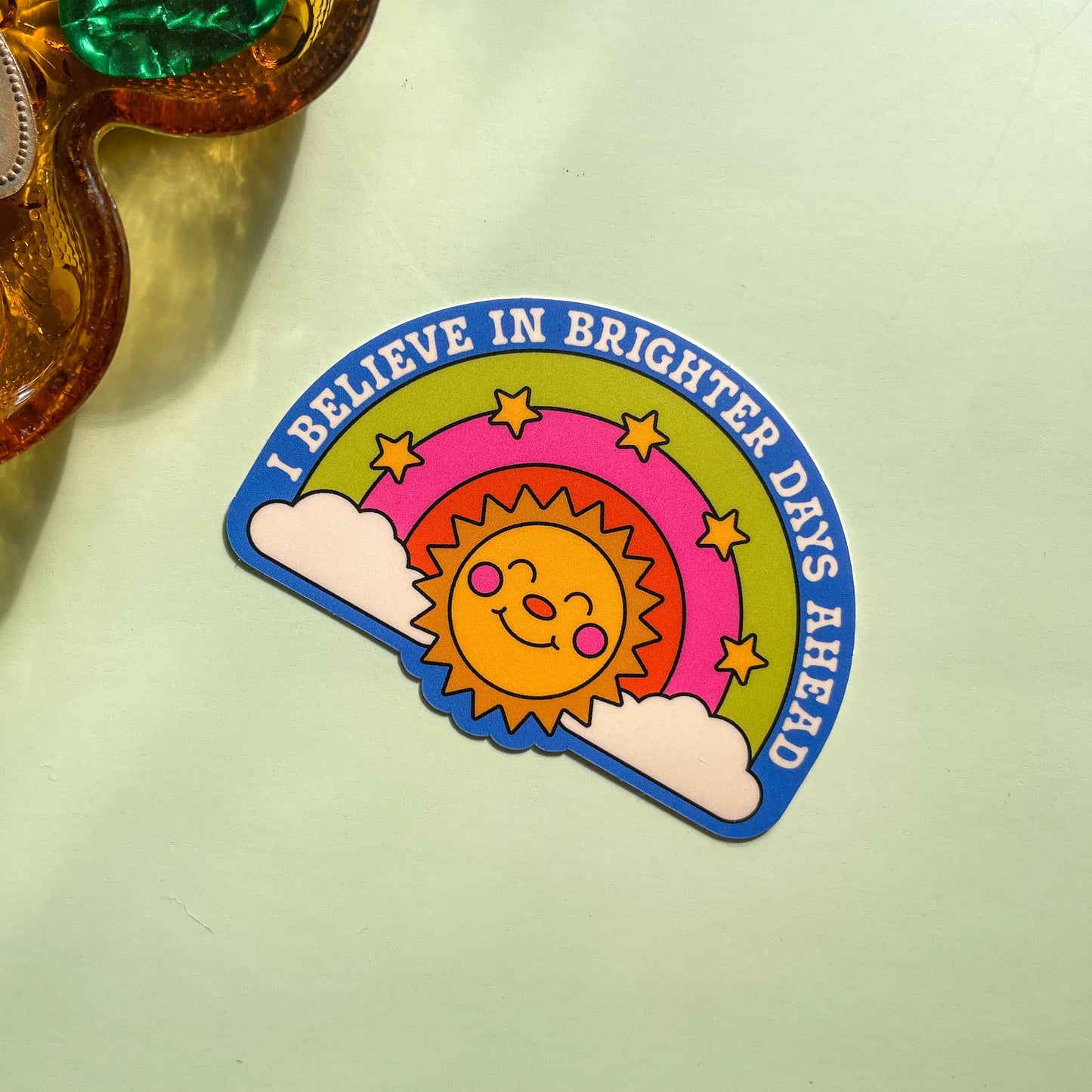 Sticker with sun design and text 'I believe in brighter days ahead' on a light green background