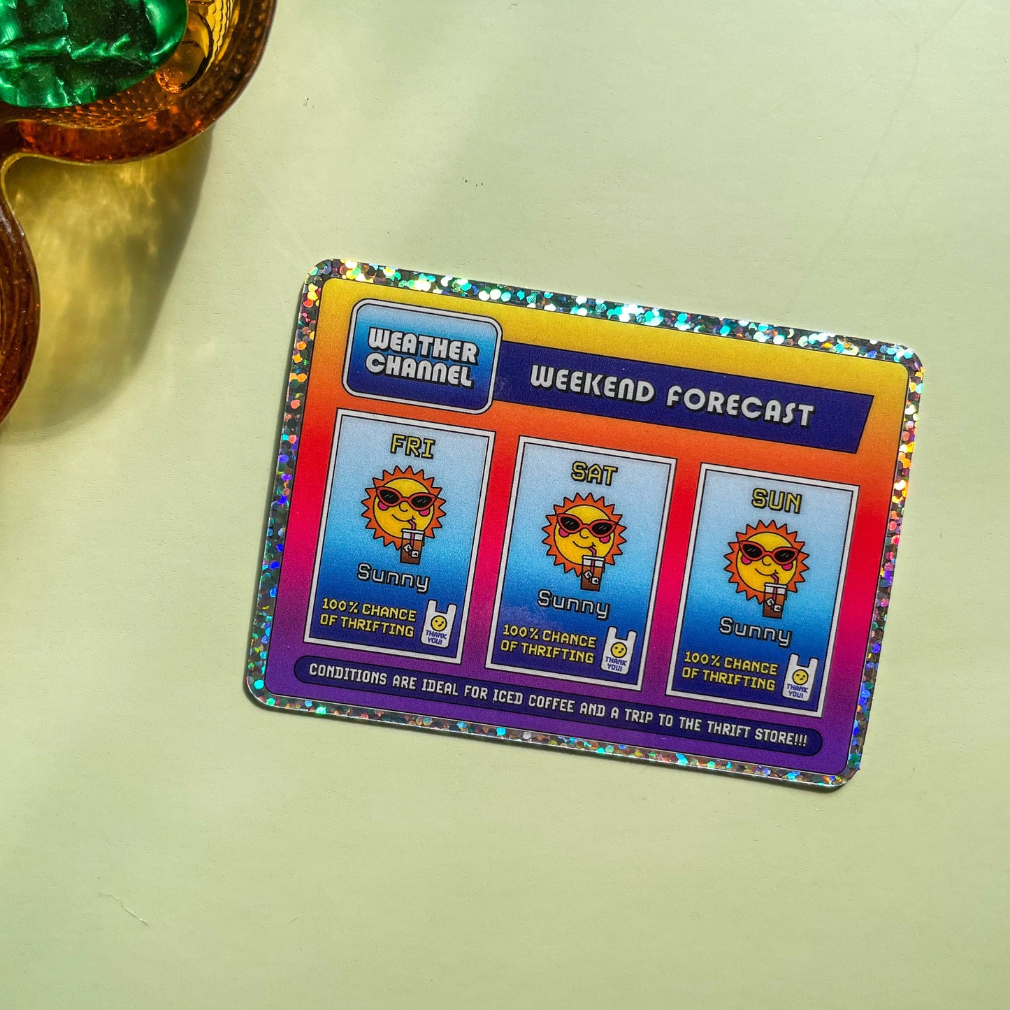 Rectangle sticker with a glitter border and a nostalgic and playful weather forecast illustration predicting sunny days ahead filled with iced coffee and good chances of thrifting on a light green background