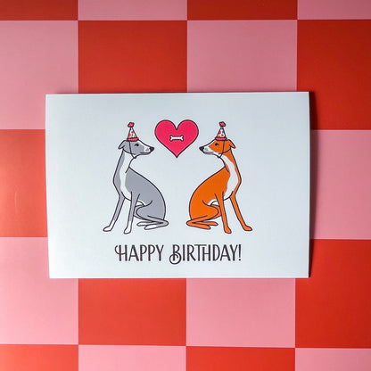 Birthday card with two italian greyhound dogs wearing party hats facing each other and a heart  with a bone in the middle and happy birthday! text at the bottom of the card on top of a red and pink checkered background