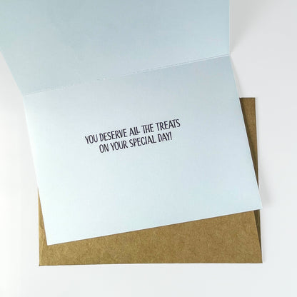 Inside of greeting card with text 'You deserve all the treats on your special day!' on top of a brown envelope and a white background.