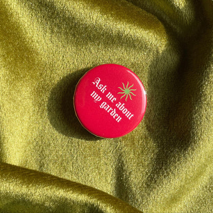 Red button that looks like a tomato with text 'Ask Me About My Garden' on a green fabric background