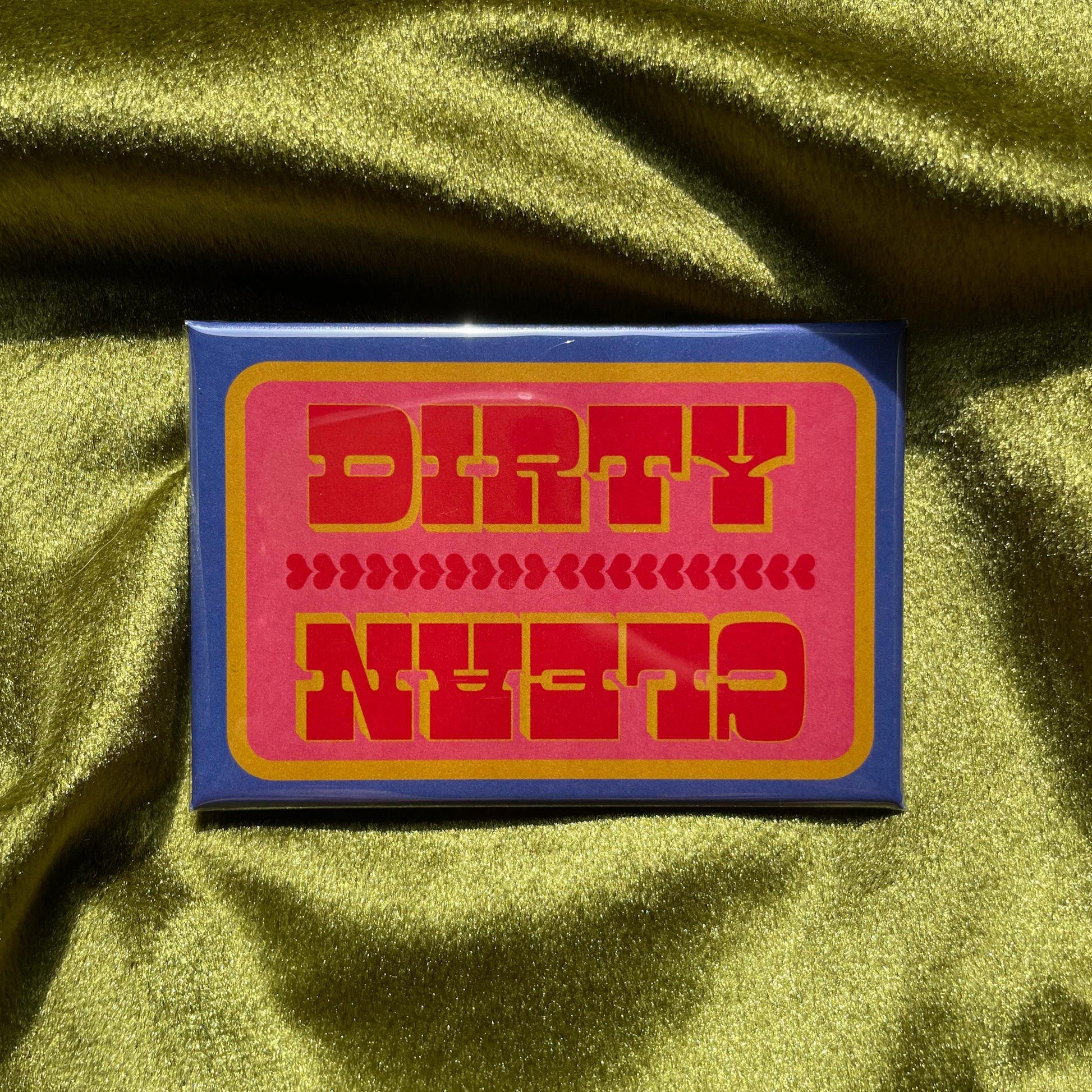A rectangular magnet with a blue and yellow border and a pink interior. The words 'DIRTY' and 'CLEAN' are in red, bold, capital letters. Between the words are rows of red hearts. The magnet is on a green, textured surface.