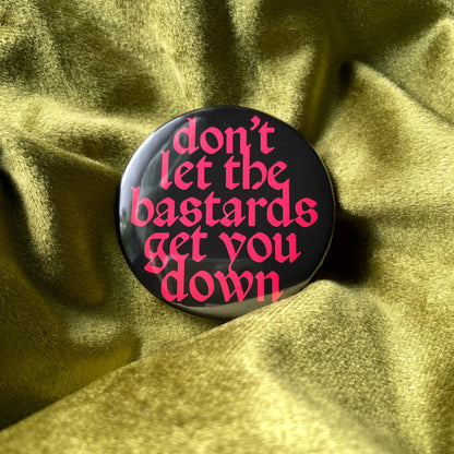A round black button with the words 'don't let the bastards get you down' in a bright pink, gothic-style font. The button is centered on a textured, olive-green fabric, creating a contrast between the dark button and the lighter background.