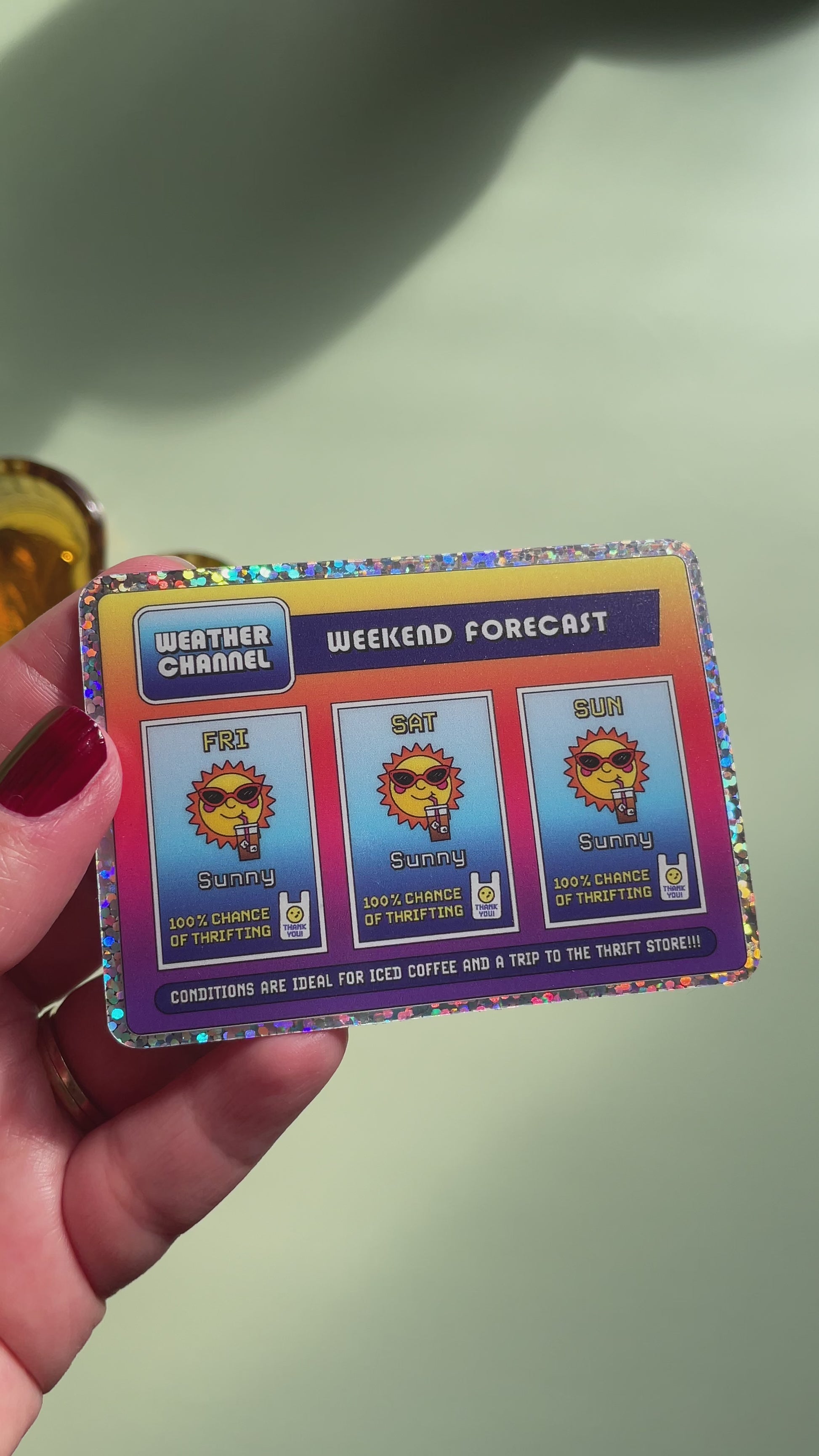 video of a hand holding a rectangle sticker with a glitter border and a nostalgic and playful weather forecast illustration predicting sunny days ahead filled with iced coffee and good chances of thrifting on a light green background