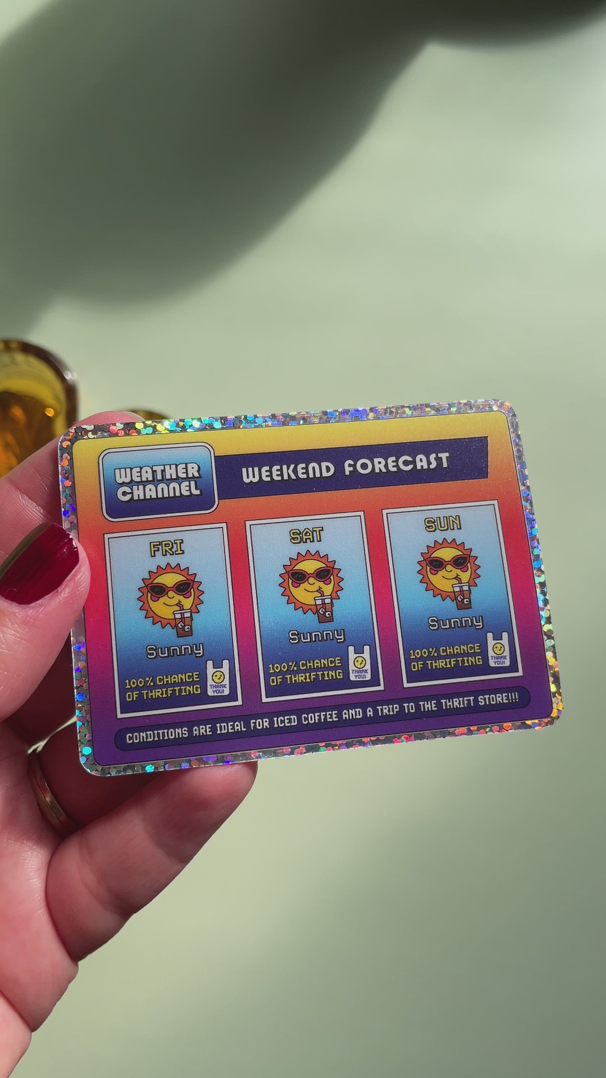 video of a hand holding a rectangle sticker with a glitter border and a nostalgic and playful weather forecast illustration predicting sunny days ahead filled with iced coffee and good chances of thrifting on a light green background