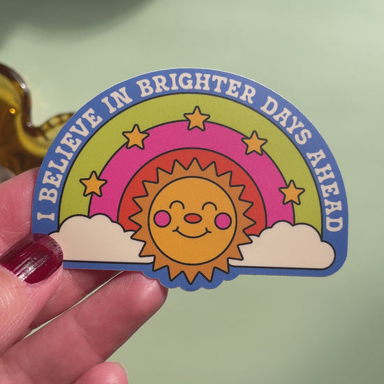 video of a hand holding a sticker on top of a light green background featuring a smiling sun below a three color rainbow with stars and the text ' i believe in brighter days ahead'