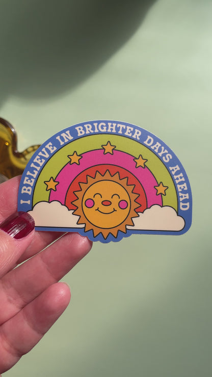 video of a hand holding a sticker on top of a light green background featuring a smiling sun below a three color rainbow with stars and the text ' i believe in brighter days ahead'
